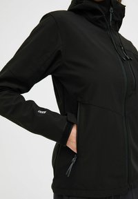 Juoda softshell striukė su užtrauktuku priekyje, reguliuojamu gobtuvu, šoninėmis kišenėmis ir Velcro rankogaliais. Lygus paviršius su prigludusia forma.