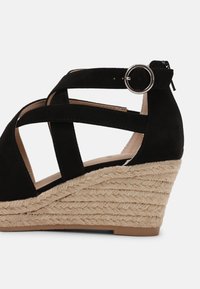 Sandale compensée en suède noir avec des brides croisées et un détail de boucle ronde. La semelle compensée est recouverte de jute texturé.