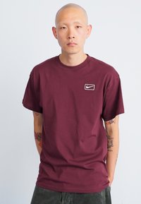 T-shirt de algodão bordeaux com decote redondo e mangas curtas; apresenta um logotipo branco da Nike no lado esquerdo do peito. Textura suave, ajuste padrão.