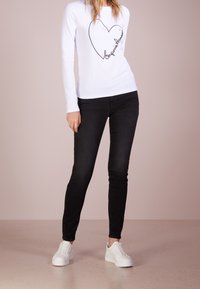 Vit långärmad skjorta med en svart hjärt-grafik och text, kombinerad med svarta slim-fit jeans och vita sneakers, stående på en ljus yta.