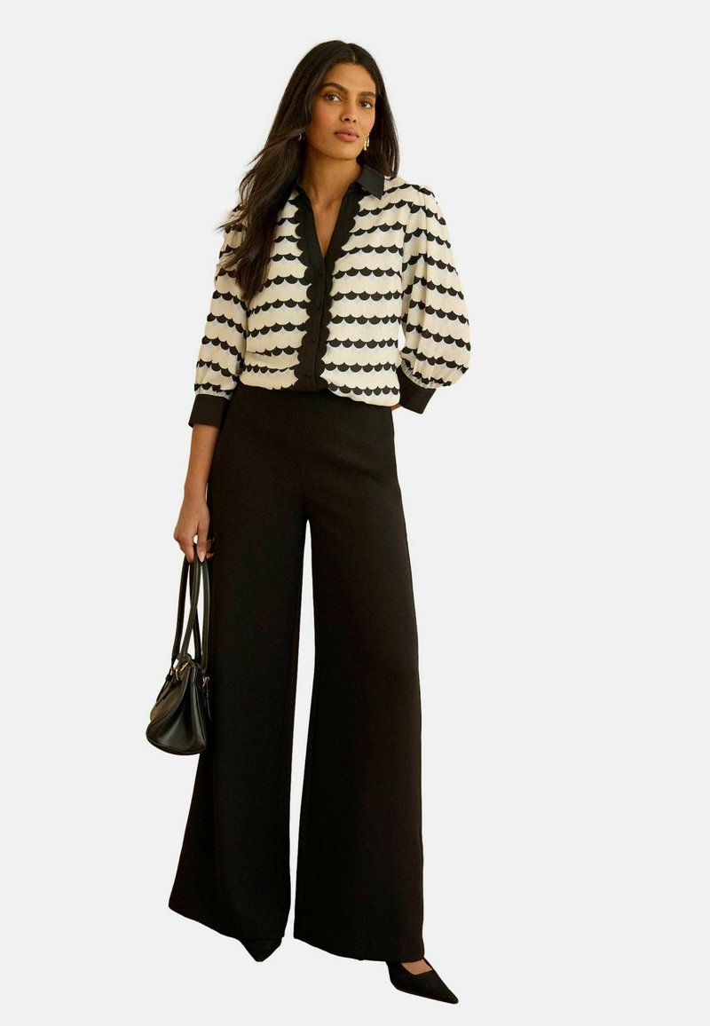 Žena u crnim širokim hlačama i bijeloj bluzi s crnim valovitim prugama, držeći crnu torbu, stoji na bijeloj pozadini.