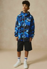 Giacca a zip con cappuccio blu e nera con design astratti; pantaloni shorts neri; sneakers bianche con dettagli grigi; calzini bianchi con strisce blu.