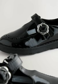 Chaussures noires en cuir verni avec un bout arrondi, une sangle élastique et un accent en métal argenté en forme de fleur. La semelle est dotée d'un motif de grip texturé.