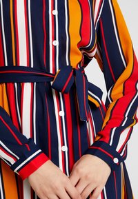 Robe chemise rayée avec des rayures verticales navy, rouge, jaune et blanche. Elle dispose d'une ceinture à la taille et de longues manches avec des poignets contrastés.
