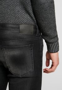 Mustat farkut, joissa on vartalonmyötäinen leikkaus, nahkainen merkki vyötäröllä, ja teksturoitu harmaa neulepusero, jossa on resorireunus hihoissa.