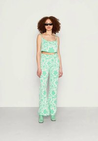 Mulher com cabelo cacheado vestindo um top cropped floral em verde-menta, calças combinando, botas mint e óculos escuros pretos, pousando contra uma parede branca.