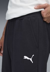 Zwarte fleece joggers met een wit Puma-logo, een elastische tailleband, zijzakken en een zachte textuur. Close-up van de broek en hand.