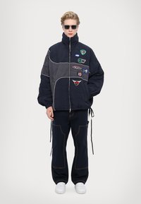 Padded navy jacket with gray sections, front zipper, multiple patches, and drawstring hem, paired with navy pants and white sneakers.