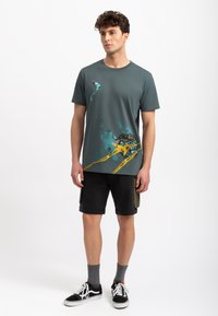 Groene katoenen T-shirt met een verfspetters grafiek, voorzien van gele "nat verf" tape. Gecombineerd met zwarte cargo shorts en zwart-witte sneakers.