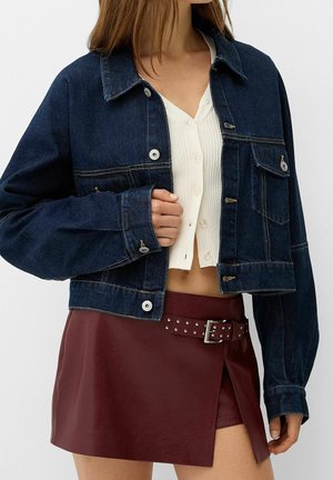 Kvinde iført en mørkeblå denimjakke over en hvid knappet croptop, kombineret med en bordeaux rød læder mininederdel med nittebælte.