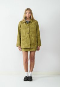 Oliwkowy zamszowy płaszcz z dwiema przednimi kieszeniami, zapięcie na guziki i dopasowaną spódnicą. Model nosi białe skarpetki i czarne mokasyny.