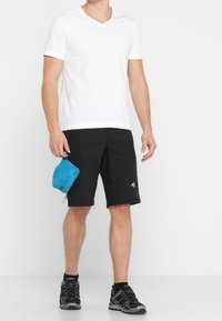 Hvid v-hals t-shirt, sorte shorts med sidelommer og sorte sportssko. En lille blå pung er fastgjort til shortsene.