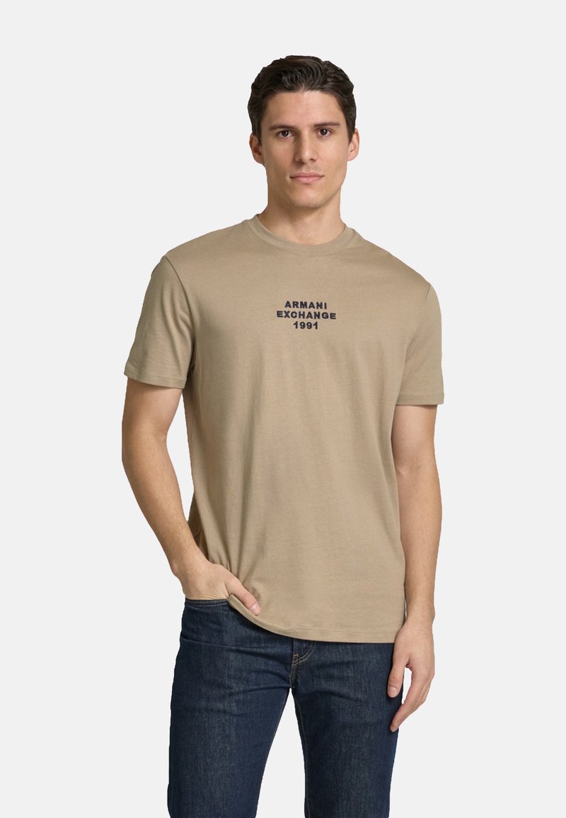 Tan bomulds t-shirt med korte ærmer, der har teksten "ARMANI EXCHANGE 1991" i mørkeblå farve centreret på brystet. Kombineret med mørkeblå jeans.