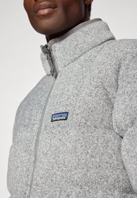 Grå fleecetröja med dragkedja och hög krage samt synliga sömmar. Har en Patagonia-logotyp på vänster bröstficka.