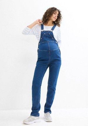 Salopette in denim blu scuro con spalline regolabili, tasche frontali e dettagli in metallo argentato. Abbinata a una camicia a righe blu chiaro.