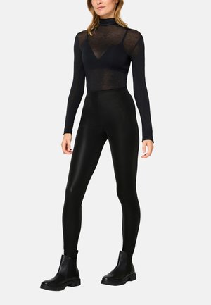 Woman wearing black sheer long-sleeve top layered with black camisole, high-waisted black leggings, and black ankle boots against plain background.