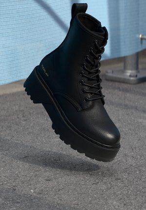 Bottine noire en matériau synthétique texturé, avec un bout rond, un laçage à l'avant et une semelle épaisse avec détail de la marque.