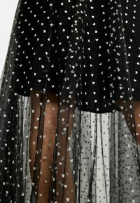 Čierne šaty s priehľadnou vrstvou, ozdobené malými striebornými srdiečkami. Tkanina má jemnú textúru a plynulý, vrstvený dizajn.