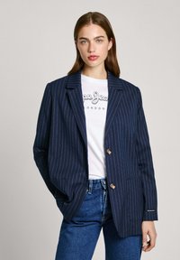 Blazer de rayas marinas con solapa con muesca, dos botones y tejido texturizado, llevado sobre una camiseta gráfica blanca y jeans azules.