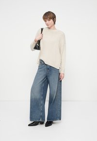 Beige pletený svetr se třásněmi po stranách, kombinovaný s širokými modrými džíny. Modelka drží malou černou kabelku.