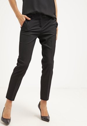 Nainen, jolla on yllään mustat, kapealahkeiset, sivutaskuilla varustetut puvuntakit, yhdistetty mustiin, teräväkärkisiin korkokenkiin ja mustaan, hihattomaan yläosaan.