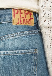 Siniset farkut, joissa on vaaleanruskea nahkalappu, jossa lukee "PEPE JEANS" punaisella värillä, ommeltu keltaisella langalla, pieni tasku näkyvissä takana.