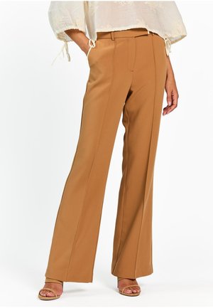 Vrouw draagt een camelkleurige, wijde, hoge taille broek, gecombineerd met beige open-toe hakken en een witte, gestructureerde blouse met gestrikte mouwen.