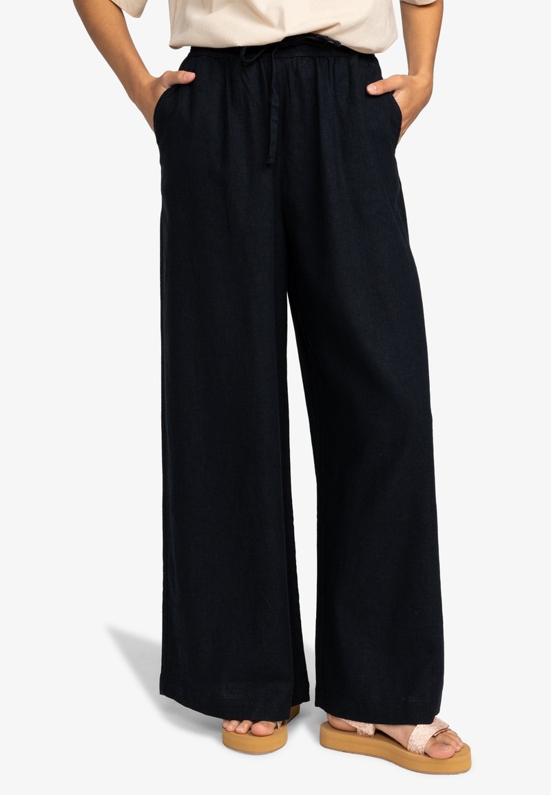 Personne portant un pantalon noir ample à jambes larges, mains dans les poches, et des sandales beiges à semelles épaisses.