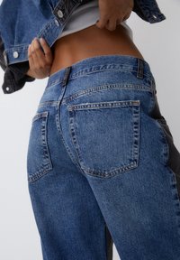 Persona levantando una chaqueta de mezclilla azul, vistiendo unos jeans azul bicolor y una blusa blanca, mostrando su abdomen contra un fondo liso.