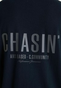 Granatowy strój z dużym napisem "CHASIN'", mniejszym napisem "AMS BASED - C.COMMUNITY" oraz pod spodem kursywą "Performance Jeanswear".