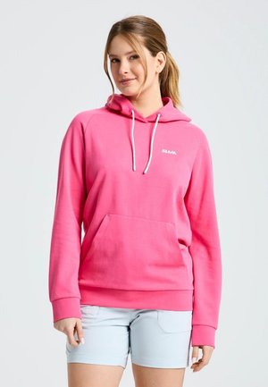Una mujer luce una sudadera con capucha de color rosa intenso, con cordones blancos, y unos pantalones cortos azul claro sobre un fondo liso y claro.