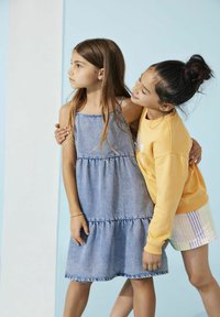 Divas meitenes valkā neformālas drēbes: viena ir ģērbusies gaiši zilā denima kleitā ar kārtām, bet otra meitene - dzeltenā sporta krekliņā un svītrainos šortos.