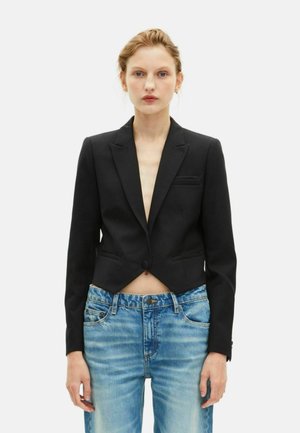 Vrouw in een zwart cropped colbert met één knoop, gecombineerd met hooggetailleerde vervaagde blauwe jeans, staande tegen een effen achtergrond.