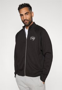 Άνδρας με σκούρα μαλλιά και γενειάδα, φορώντας μαύρο φερμουάρ μπουφάν με κείμενο "City" και ανοιχτό γκρι παντελόνι, στέκεται μπροστά σε απλή φωτεινή φόντο.