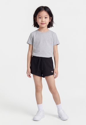 Mladé dievča stojace čelom dopredu, oblečené v sivom tričku, čiernych šortkách so sťahovacou šnúrkou, bielych ponožkách a bielych teniskách na jednoduchom bielom pozadí.