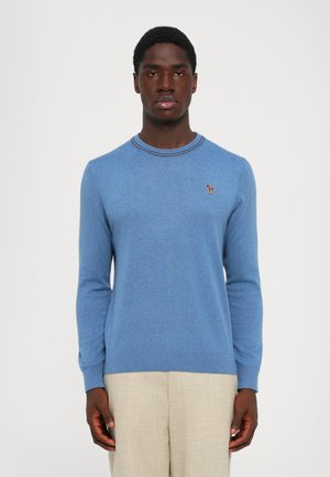 Man wearing a light blue long-sleeve sweater with a small multicolored dog emblem on the left chest, paired with beige pants.