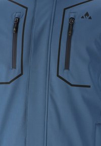 Blå mjukskaljacka med två zippered bröstfickor, svarta detaljer och en strukturerad tyg. Geometriska sömlinjer ger extra detalj.