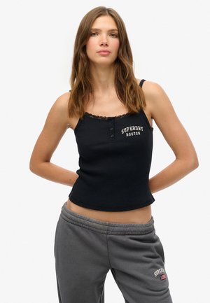 Kvinde med langt brunt hår iført sort tanktop med teksten "Superdry Boston" og grå joggingbukser, stående med hænderne bag ryggen.