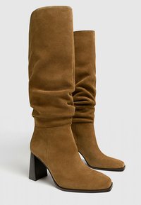 Bottes hautes à talons en daim marron avec un design décontracté, un bout carré et un talon bloc texturé. La partie supérieure tombe doucement, offrant une silhouette détendue.