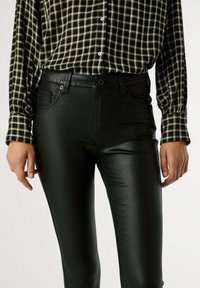 Jeans skinny de piel sintética negra con una textura suave, bolsillos frontales y trabillas, combinados con una camisa de cuadros verde y negra.
