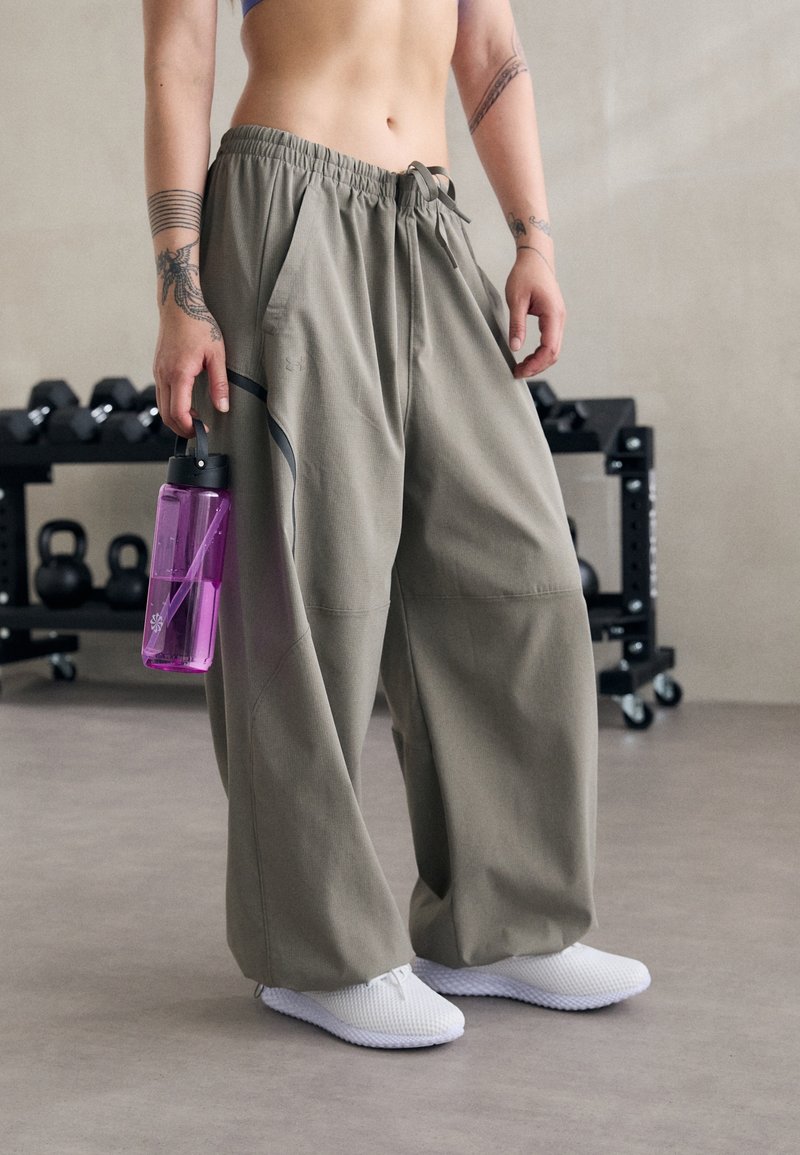 Personne en pantalon gris ample et baskets blanches tenant une bouteille d'eau violette avec des poids de gym en arrière-plan.