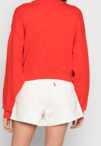 Persona che indossa una camicia rossa a maniche lunghe e pantaloncini di jeans bianchi, mostrata di spalle su uno sfondo semplice.