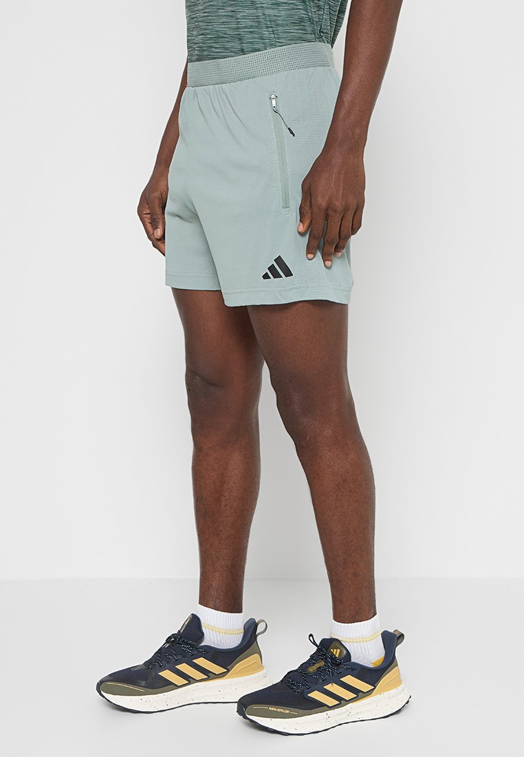 Pantalones cortos deportivos de color gris claro con una cinturilla texturizada, bolsillo lateral con cremallera y logotipo negro de Adidas. Combinados con zapatillas oscuras y calcetines blancos.