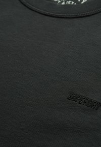 Primer plano de una tela negra con el logotipo bordado en negro de "SUPERDRY" cerca de la esquina inferior derecha, debajo de un cuello redondo.
