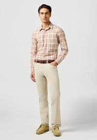 Camisa beige a cuadros con acentos rojos, pantalones de color claro y zapatos tipo mocasín amarillos; el modelo está de pie con una mano en el bolsillo.