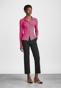 Cardigan ajusté en rose côtelé avec col et fermeture à boutons, associé à un pantalon noir ajusté. Modèle portant des talons bicolores.