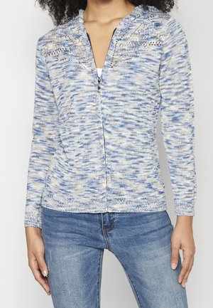 Femme portant un pull en maille zippé marbré bleu et blanc à manches longues et un jean bleu, debout les mains détendues sur les côtés.