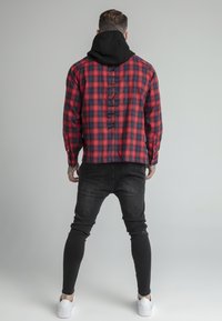 Camicia di flanella a quadri rossi e neri con cappuccio nero, caratterizzata da un design unico sul retro e jeans neri slim-fit, abbinati a sneaker bianche.