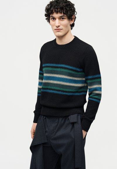 Jeune homme aux cheveux bouclés portant un pull noir à rayures horizontales bleues, vertes et grises, ainsi qu'un pantalon à fines rayures avec un détail en tissu noué.
