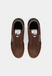 Biezi brūni zamšādas kedas ar gumijas zoli, melniem akcentiem un polsterētu mēli. Satur Reebok zīmolu un apaļas šņores.